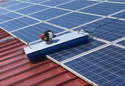Silver steel solar cleaning robot with blue rotating brush cleaning solar panels on a red-colored rooftop