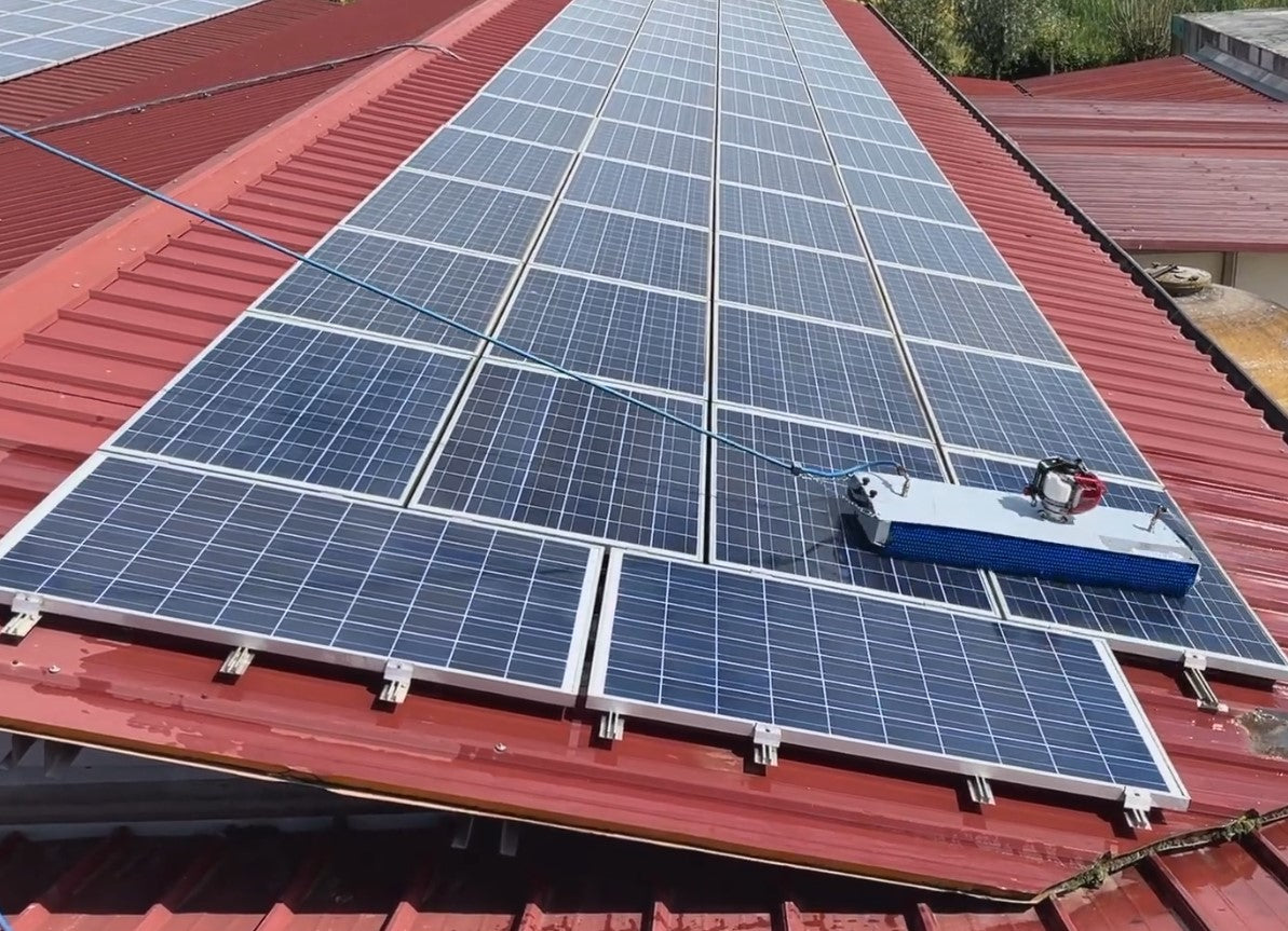 Silver steel solar cleaning robot with blue rotating brush cleaning solar panels on a red-colored rooftop