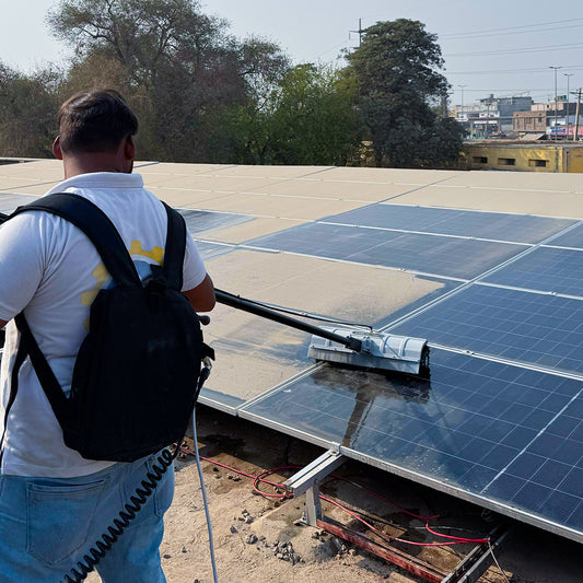 Man using a long black-handled solar panel cleaner with a silver steel rotating brush to scrub panel surface