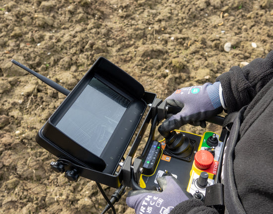 Man wearing gloves operating a remote control device with a screen and signal antenna, likely controlling machinery remotely.