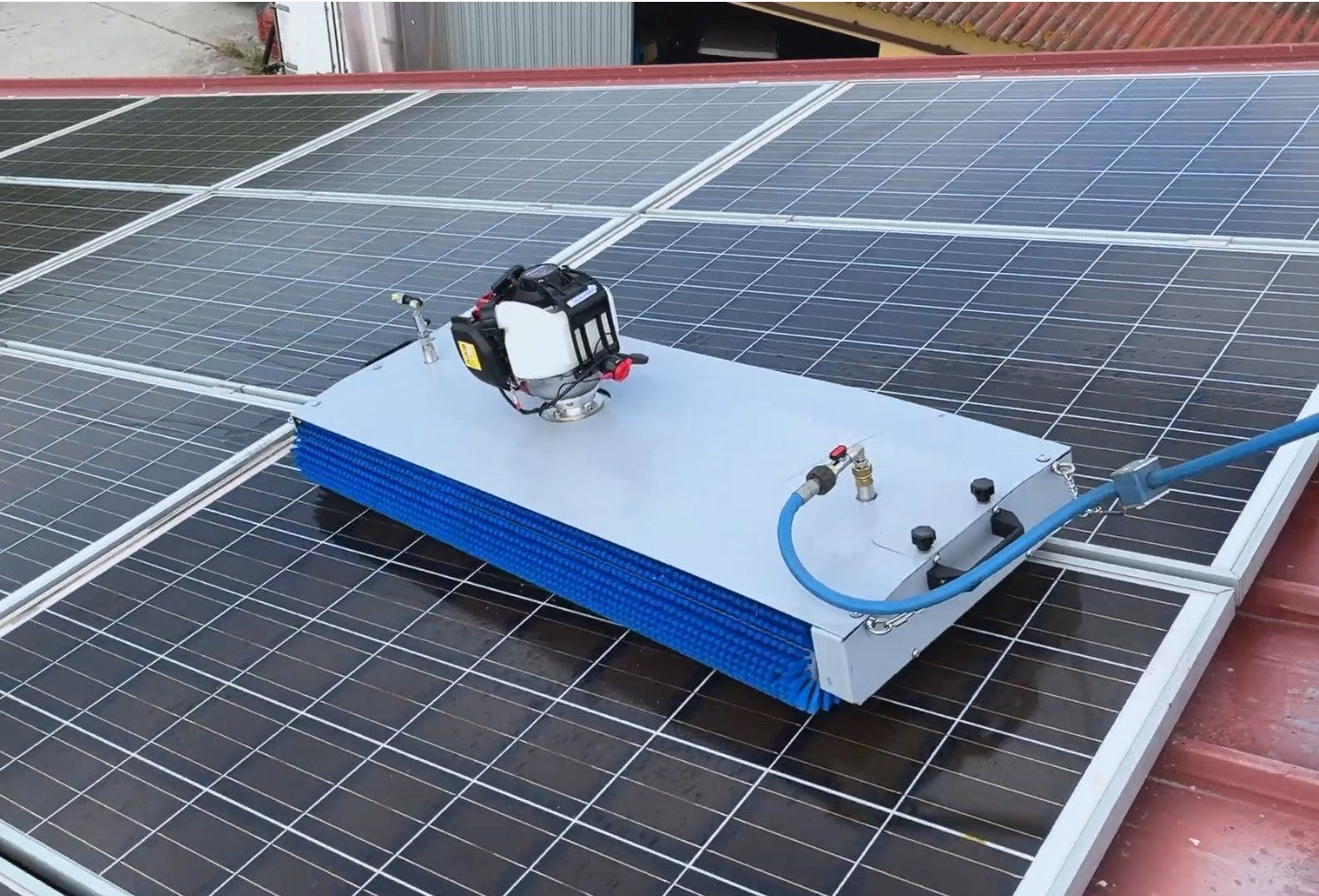 A solar panel cleaning machine mounted on a roof, with blue hoses and machinery visible against the solar panels.