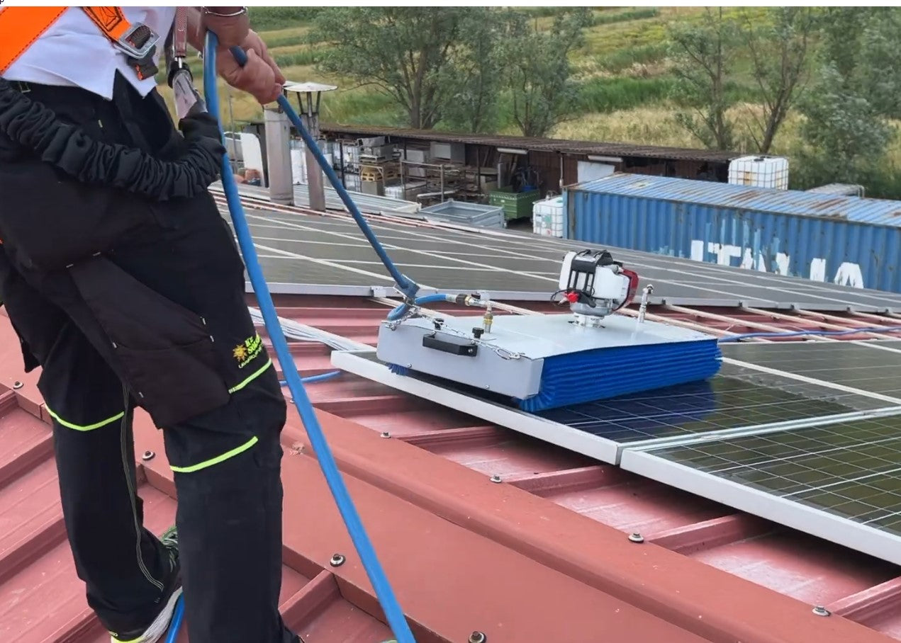Man cleaning solar panels on a red roof using a silver metal solar cleaning robot with blue rotating brushes