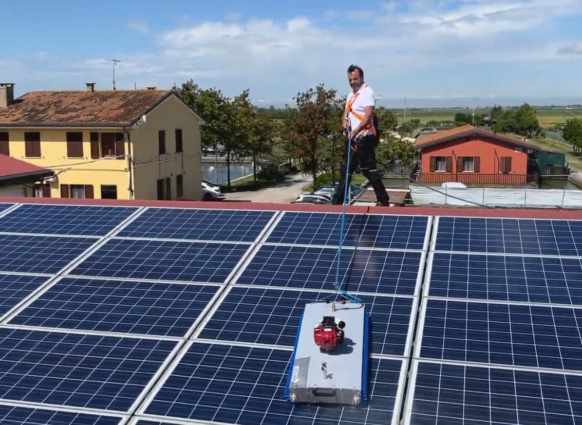 Man using a silver steel solar cleaning robot with blue rotating brush to clean solar panels on a rooftop.